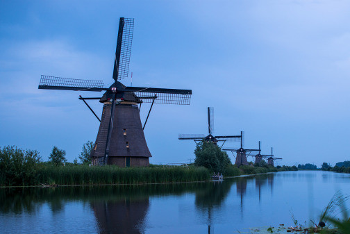 Photo from the Netherlands showing at least five historic windmills lined up along a watercourse and reflected in the water. (Photo: R Boed/Flickr (CC BY 2.0 DEED).) 