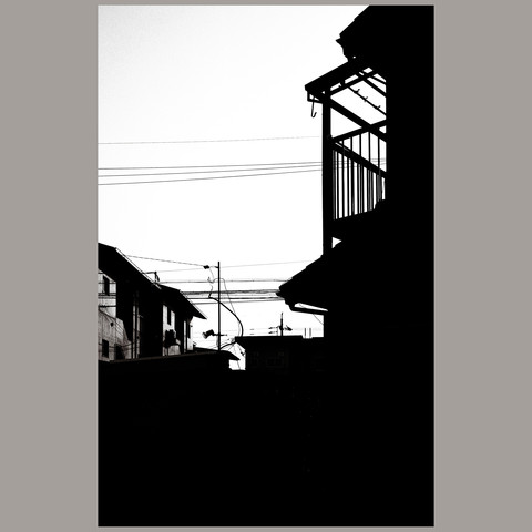 This photograph captures a typical afternoon in a Nagoya neighborhood in high contrast monochrome. Buildings, both residential and utility, are defined by their sharp silhouettes against a bright sky, punctuated by a network of power lines. In the center, a single spotlight attached to a pole stands out, a sentinel in the urban tapestry. The play of light and shadow creates a dramatic and yet commonplace snapshot of urban Japan.