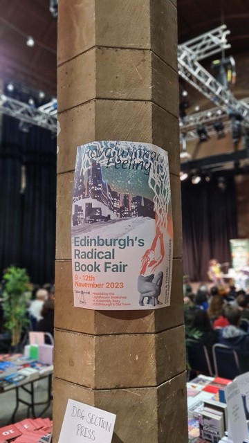 Poster for Edinburgh’s Radical book Fair displaying event information, white paper with green and orange text, the illustration is of a woman, lower half photo, of legs, upper half illustrated long hair, arms aloft fanning into spell/flames roots that arc up left sideover top of poster, in background a stylised nighttime street and city scape.  The poster is in the middle of a large stone pillar, to the side and background out of focus, tables of books, spectators, ceiling truss, speakers and interviewers seated on stage, some plants and joyful activity.