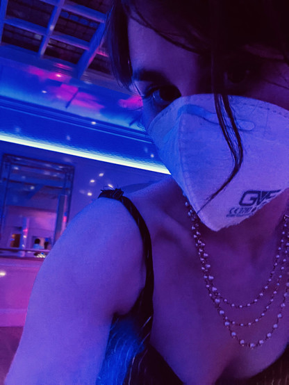 A woman wearing a strappy dress, faux pearl necklace and N99 mask under purple UV style light