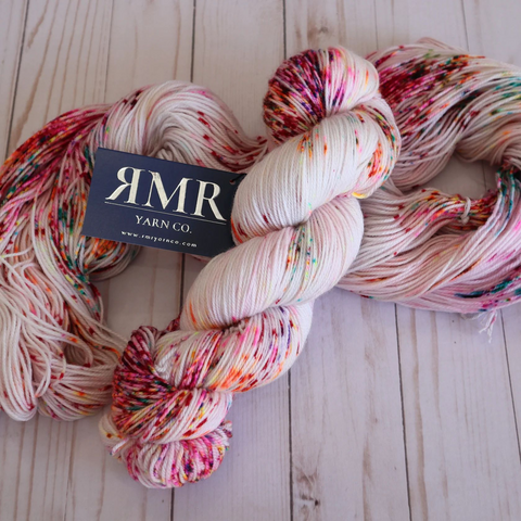 Pale pink yarn with dark and bright speckles in red, pink, orange, green etc. One skein is shown twisted, the other untwisted on a light wooden background.