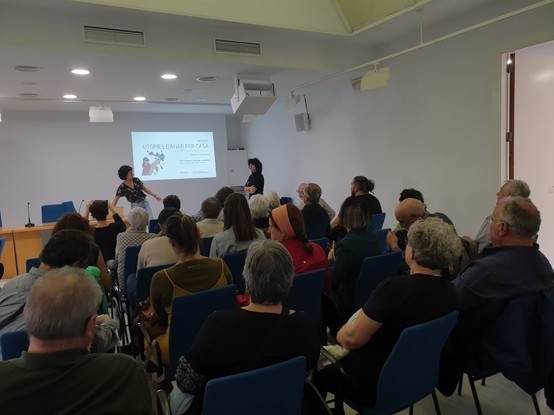 Foto de las personas asistentes sentadas en sillas y las ponentes de la jornada junto a la proyección.