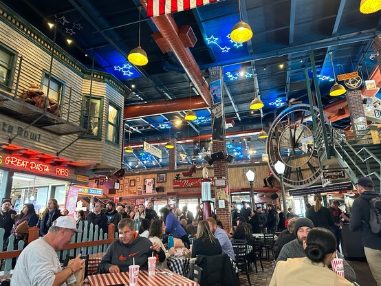 Inside of a restaurant that has two floors. It is made to look a bit like you are outside on the street. There are neon blue star lights on the ceiling. There is a like 20 foot black analog clock hanging by the staircase on the right. The tables have red and white gingham tablecloths. 