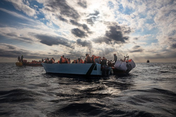 Ein großes hellblaues Holzboot auf dem Meer. An Bord viele Personen mit Rettungswesten.
An der Steuerbordseite hat ein RHIB angedockt, das gerade Menschen an Bord nimmt. Ein weiteres RHIB sichert ab.
Im Hintergrund ist ein großes Schiff zu sehen.