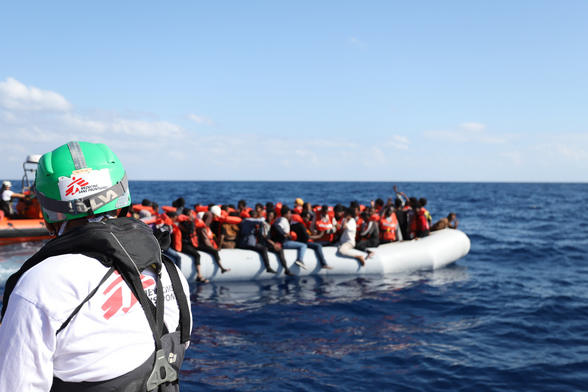 Auf einem Schlauchboot dicht gedrängt Menschen, die teilweise auf den Schläuchen sitzen. Viele tragen Rettungswesten, aber einige noch nicht. Im Hintergrund ist ein RHIB zu sehen. Im Vordergrund ist von hinten ein*e Seenotretter*in zu sehen.