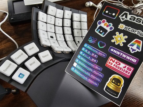 The right hand part of my ergonomic split keyboard with an Astro sticker sheet on top of it. One thumb key on the keyboard has a very tiny Astro logo that I have cut out of another sticker on the sticker sheet.