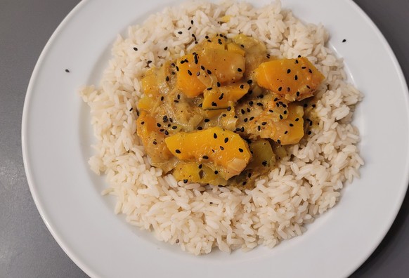 Farbfoto: Auf einem weißen Teller liegt eine Portion Reis, in der Mitte eine Portion orangenes Curry aus Kürbis, bestreut mit etwas schwarzem Sesam. 