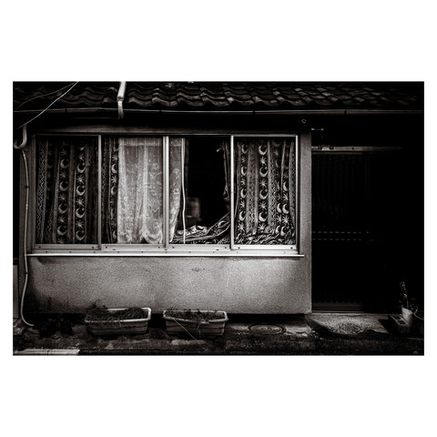 A monochrome image captures a moment of quiet desolation in Nagoya. An air conditioner, just out of frame to the left, suggests the presence of life, but what dominates the scene are signs of neglect. Lace curtains, stained and frayed, hang behind a pair of glass windows adorned with ornate grilles, contrasting with the building's dilapidated exterior. Below the windows, two planters sit with their contents wilted and lifeless, symbolizing decay amidst the urban landscape. The stark play of light and shadow highlights the textures of despair and ennui in this urban tableau. 