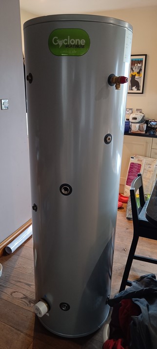 A new hot water cylinder, as tall as a person, waiting installation. It is stainless steel, wrapped in insulation then wrapped in more steel.