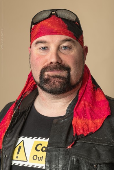 Matt Whifield, actor, as "Stallion", the British wrestling commentator and personality. He is wearing a red bandana, black vest top and black biker kutte. He has a black beard and piercing blue eyes.