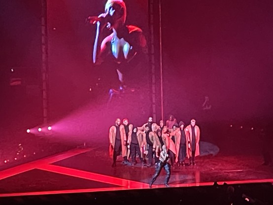 Doja Cat on stage with her dancers. Behind the stage is a huge floor to ceiling monitor showing a closeup of Ms Cat singing.