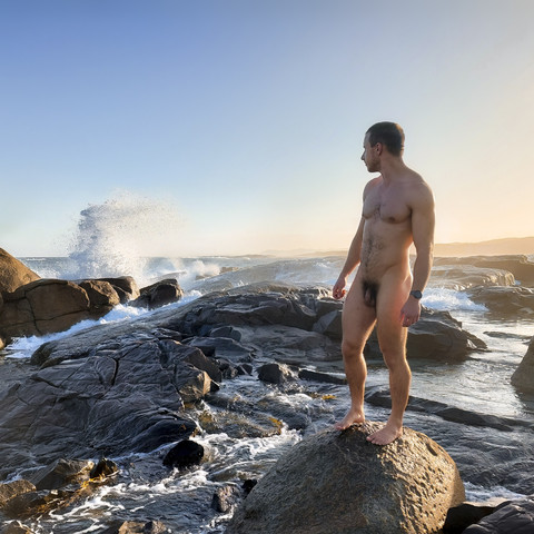 A naked man stands on a rock by the sea, with waves crashing over rocks in the background.