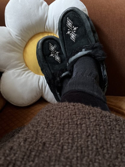 Feet wearing black moccasins (with beading on upper of 2diamond shapes) resting on a pillow shaped like a daisy 