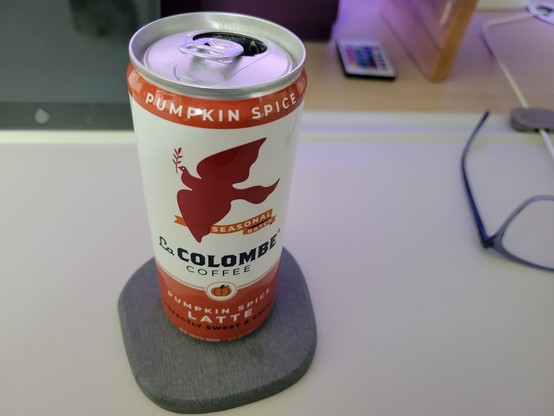9 fluid ounce can of La Colombe Pumpkin Spice Latte Coffee, on a gray diatomaceous coaster on an off-white desk mat on a maple desk. 

The La Colombe can has a white background with orange borders at top & bottom, and a dark orange silhouette of a dove of peace in flight while holding an olive branch, flying above a banner reading "Seasonal Batch". Below this is a small pumpkin enclosed in a gold circle with a white background. Other text on the can: "Perfectly Sweet & Creamy".