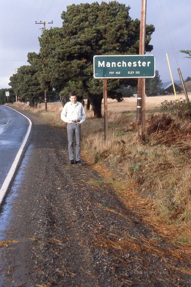 Sign for Manchester, pop 462 elev 120. A man stood next to it facing the camera.