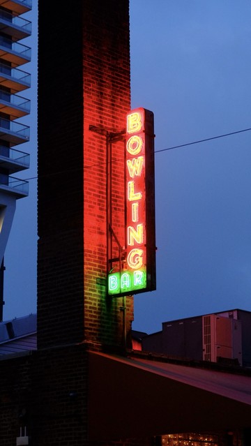 A red neon sign for a bowling alley attached to a brick chimney with an apartment high rise in the background