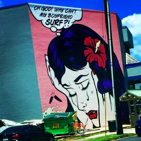 A large wall mural depicting a comic of a crying woman with red hibiscus in her black hair. There is a thought bubble above her head exclaiming "Oh God! why can't my boyfriend SURF?!