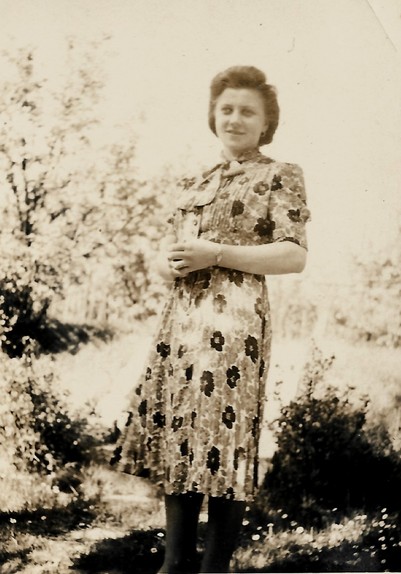 black and white photo dated 1941 of a young woman in a dress with flowery pattern, standing outdoors with some shrubs and grassland in the background. Clearly posing to show off the dress, as there are other photos from the same date with different outfits. 