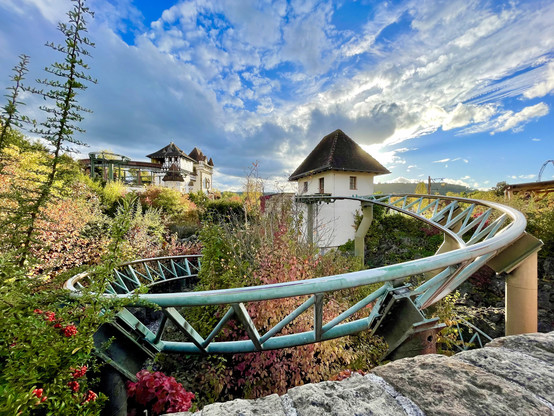 Final helix of G'Sengte Sau roller coaster. Plenty of colourful shrubbery on the ground, a couple of buildings in the background, and bright blue cloudy skies.