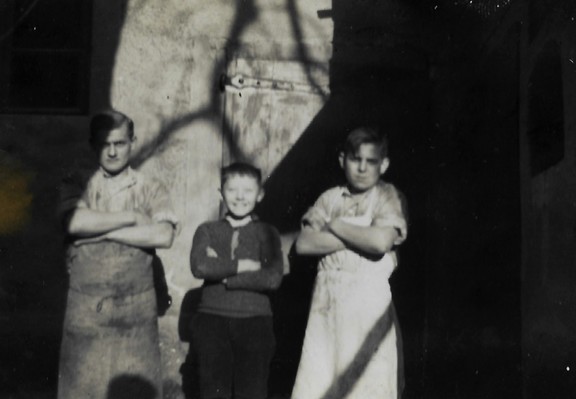 black and white photo from the 1930s showing a young man and two boys posing with their arms crossed. The younger boy stands in the middle and grins, while the other two look more fierce. The background is a building with dramatic shadows of a bare tree projected onto it by a wintry low sun. 