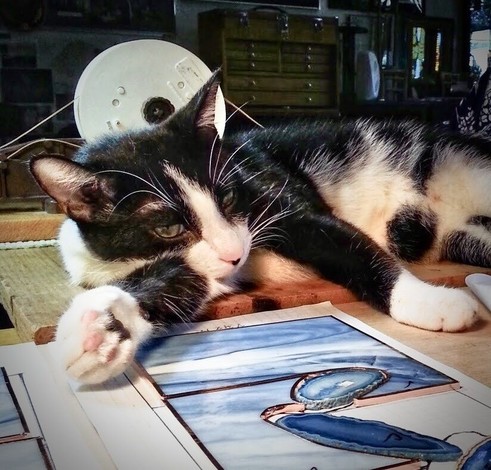 A black and white cat lounges on a table with its paw resting on a stained glass project in process. 