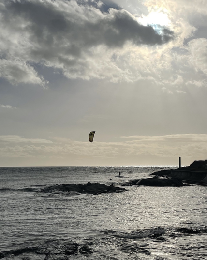 Kite-surfer on waves, bright sun peeking behind clouds with rocky shore to the right of frame