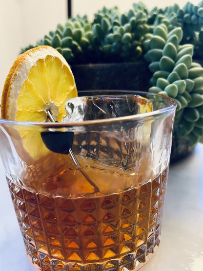 An Old Fashioned cocktail in a faceted rocks glass with a large ice cube — garnished with a cherry and dehydrated orange slice.
