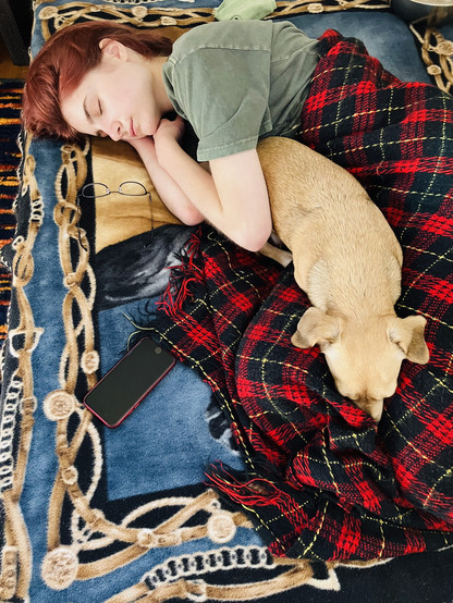 A (currently) flaming red-headed teen girl rests curled around her emotional-support pup, Oona on patterned blankets of red plaid and blue and beige. 