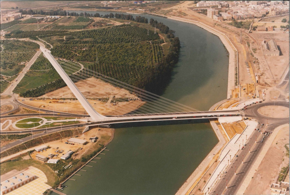 an aerial photo of the bridge , a river underneath it brown land and sections of green farms nearby 