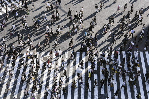 aerial photo of a street intersection with several large zebra crossings and more than 100 pedestrians casting long shadows. 