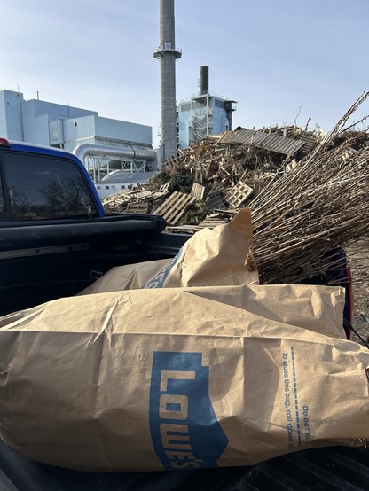 Photo of the back of a pickup truck with some yard waste bags with sticks sticking out. In the background is a pile of wood and a large building with a smokestack. 