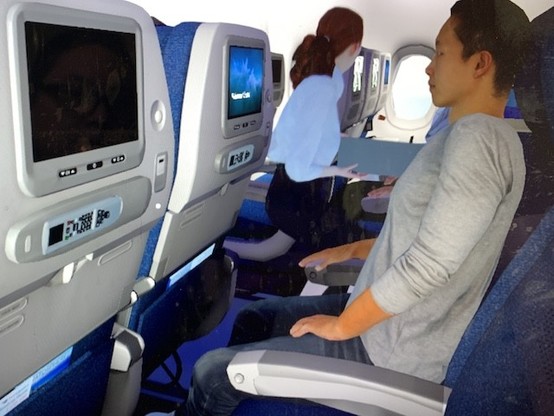 Screen shot of a simulated row of airliner passenger seating, with a young Southeast Asian-looking man in the aisle seat. 