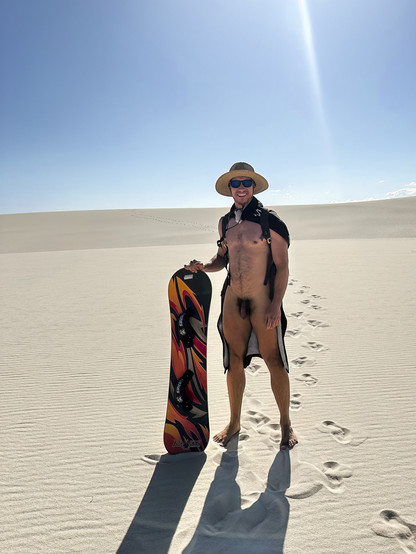 A naked man with a hat, sunglasses, cape and backpack standing in a desert with his hand resting on a sandboard that is planted in the ground.