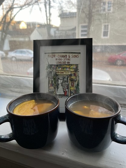 Two cups of mulled cider with orange slices on a window sill with a view of a quiet city street. 