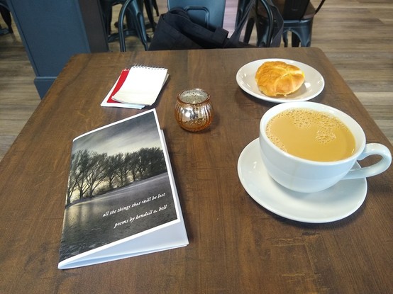 On a dark square wood table, clockwise: sausage egg and cheese croissant on white ceramic plate; large white ceramic cup and saucer with coffee; Kendall a. Bell's chapbook "all the things that will be lost"; blank memo pad on top of a red memo pad; and amber votive candle.