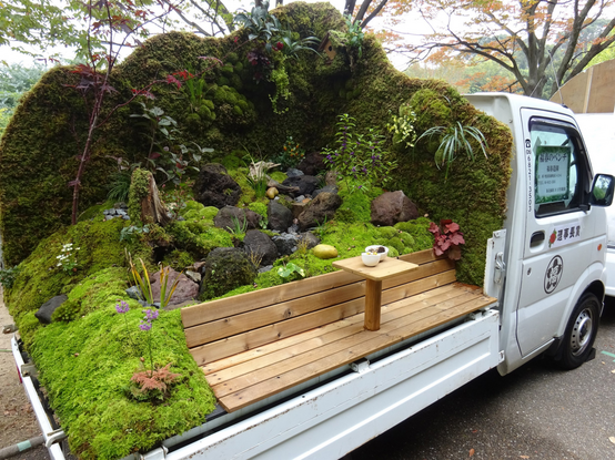 photo of a white kei truck with a beautiful garden built on it. There is a wooden bench with a small tea table built into the side of the truck. The bench is very inviting and there is a cup of green tea  sitting on the table. 
