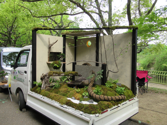 A white kei truck with a garden scene built onto the truckbed. It is filled with grass and moss and various plants to recreate the feel of a garden. There are also some additional items added, like an old, thick rope, some aged pots, and a bonsai tree.