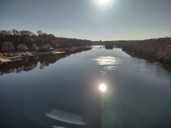 Photo: View from midspan of the Strawberry Mansion Bridge, over the Schuylkill River, Philadelphia. On the left (east) bank is the Lasalle boathouse. Straight ahead, on the shore at a sharp bend in the river, rises the Beaux-Arts dome of Memorial Hall (from the 1876 Centennial Exposition, now the Please Touch Museum). Both riverbanks are lined with deciduous trees, either bare of leaves or covered with the multiple hues of autumn — yellows, oranges, reds. The sky is cloudless. The sun is bright and reflected on the surface of the water.