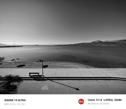 Photo en noir et blanc du bord de la mer Méditerranée. Lignes formées par la route, la plage et l'horizon. Ombres d'un arbre, d'un banc et d'un parasol. Itea, Grèce. 2023.