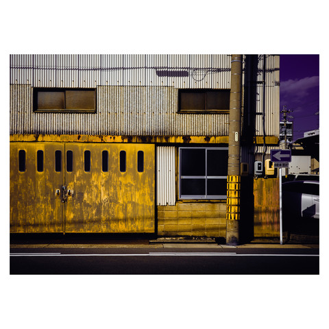 The photograph shows the side of a light industrial building painted in yellow, with signs of wear and weathering. The aged paint reveals parts of the building's structure. It's a clear day, and the building stands in a zone that blends both residential and industrial elements, with the street in the foreground and other buildings in the distance.