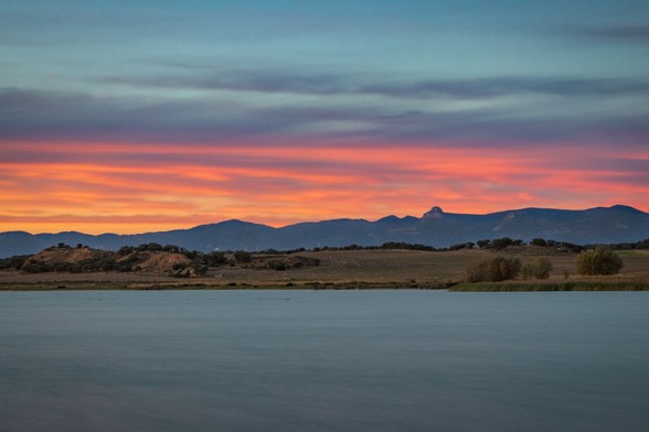 Se puede ver en primer plano un embalse de agua, después campos y montañas a lo lejos y un cielo con algo de nubes y colores del atardecer | You can see in the foreground a water reservoir, in the middle some Fields and in the background some mountains and sunset Sky.