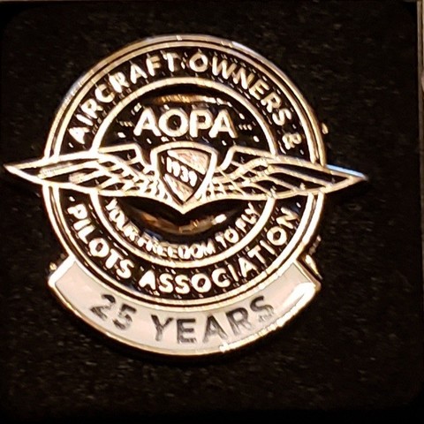 A lapel pin with a black background and outline emblem of "wings" with "AOPA" above the wings and the words, "Aircraft Owner's & Pilot's Association" around the outside. If you really squint hard, you can see the words, "Your freedom to fly" inside the circle below the wings. At the bottom of the pin is a white banner that reads, "25 years".
