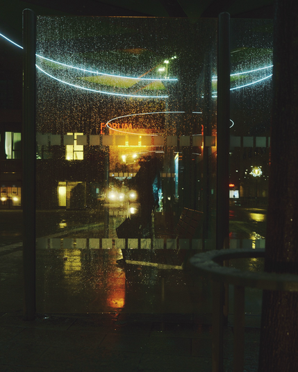 Mehr oder minder abstrakte Fotografie. Eine verschwommene Person hinter einer mit tropfen benetzten Scheibe bei Nacht mit warmen Lichtern im Hintergrund und led Lichtringen als Spiegelung 