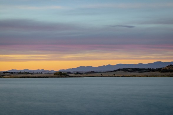 Se puede ver en primer plano un embalse de agua, después campos y montañas a lo lejos y un cielo con algo de nubes y colores del atardecer | You can see in the foreground a water reservoir, in the middle some Fields and in the background some mountains and sunset Sky.