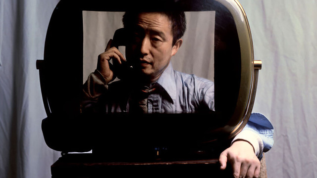 Screenshot of "Moon Is the Oldest TV" featuring Nam June Paik behind a camera monitor holding a black vintage telephone handset speaking with white curtains as a backdrop.