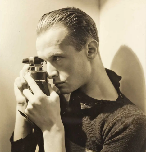 Henri Cartier-Bresson by George Hoyningen-Huene, 1935, via Museum of Modern Art, New York.

A black and white picture of a young white man with light, slickback hair. He is standing sideways, holding an old camera in front of his right eye. 