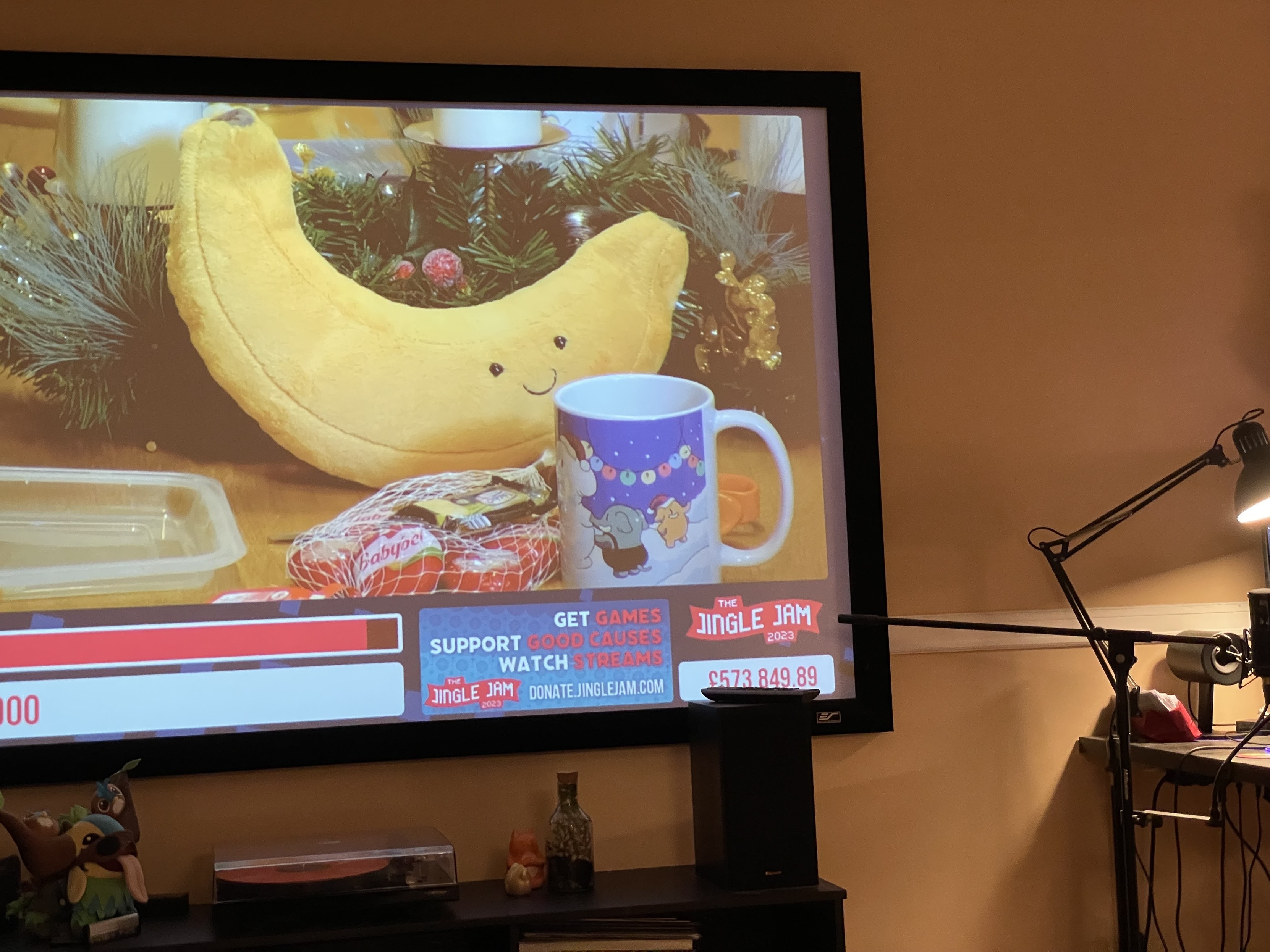 A large projector screen showing the Jingle Jam stream on Twitch. The stream is on break and showing the feed from the Yogscast office. On the coffee table, next to a large banana plushie, stands a mug featuring colorful elephants building an elephant snowman under a blurple sky. 