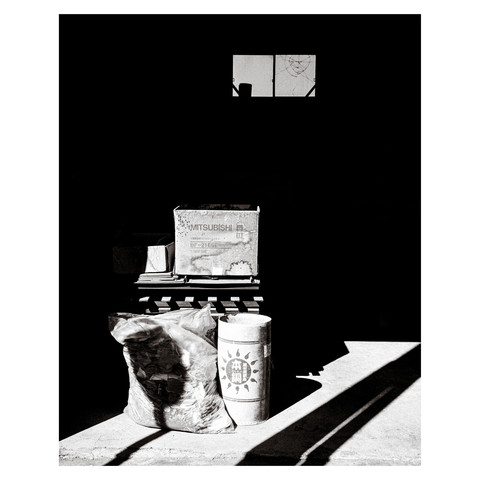 The photo is a high-contrast black and white image capturing a seemingly mundane corner outside a metalwork factory. Objects including a trash bag, wooden pallets, a cardboard tube, and a box are arranged in a visually interesting way, with the play of shadows creating a dramatic effect. The distinct shadow of a no parking sign overlays the scene, suggesting a space typically overlooked but momentarily significant.