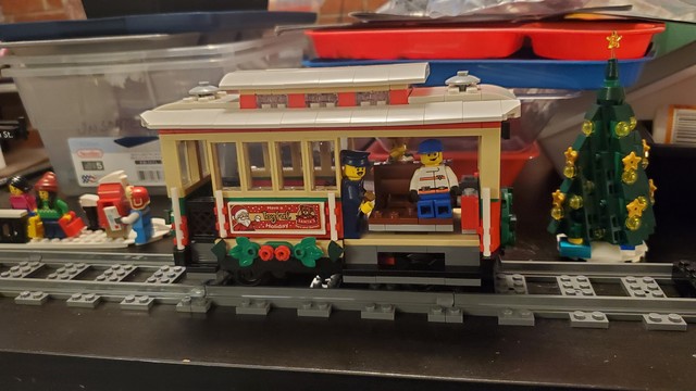 Lego trolly car on a section of track, with a Lego Christmas tree and platform with Minifigures in the background.