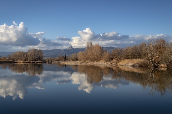 Se puede ver un pequeño embalse de agua en el cual se reflejan algunos árboles, vegetación, nubes y al fondo parte de la Sierra de Guara (Huesca, España) | You can see a little reservoir with some reflections of trees, vegetation, clouds and in the background a mountainscape.